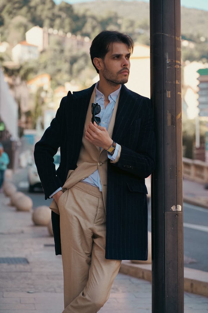 gallery-4 A fashionable man in a suit leans against a post in an urban setting, exuding confidence.