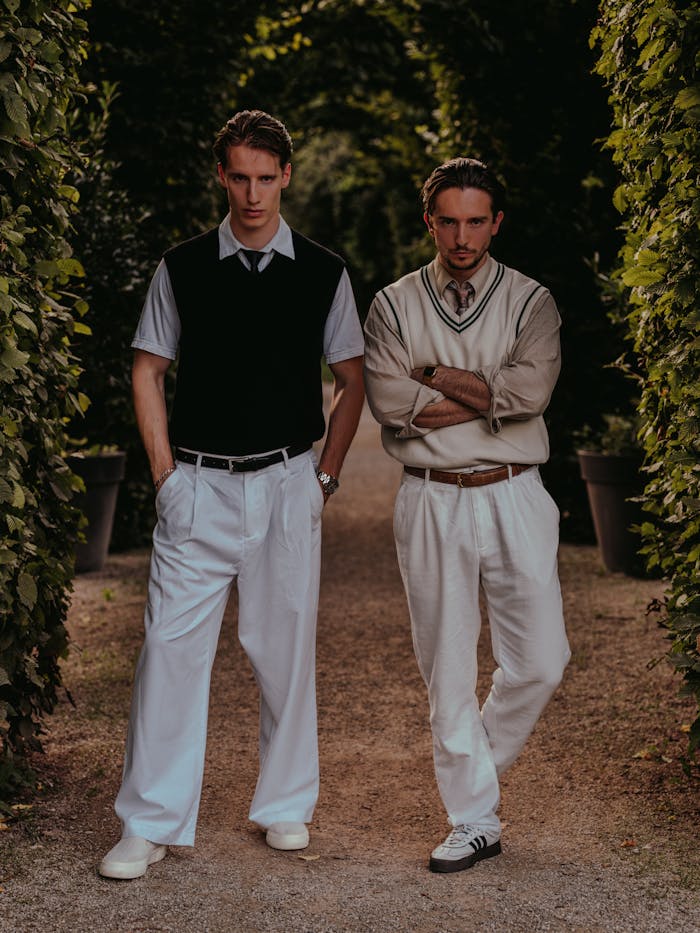gallery-2 Two men in vintage-inspired fashion pose confidently in an outdoor garden setting.